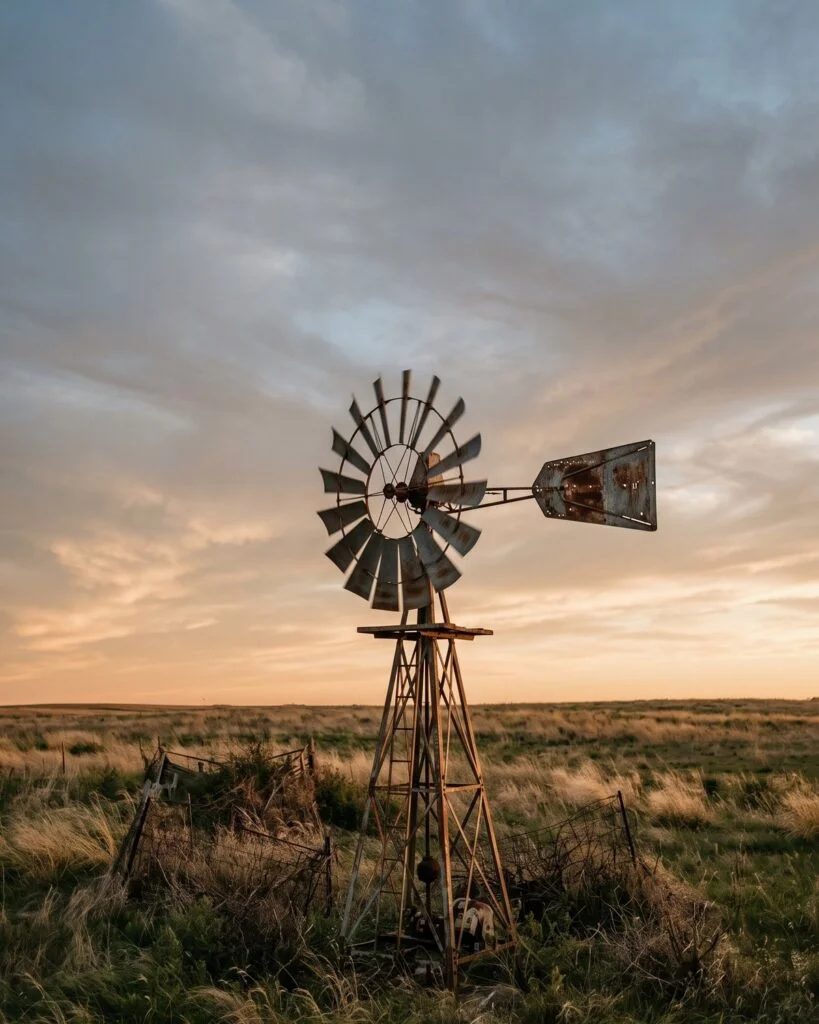 A rusted windmill in a windy field at sunset, blades turning again after a long pause.